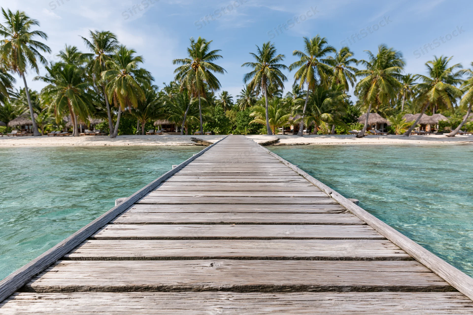 Wooden dock extending over turquoise water toward palm-fringed beach with thatched huts