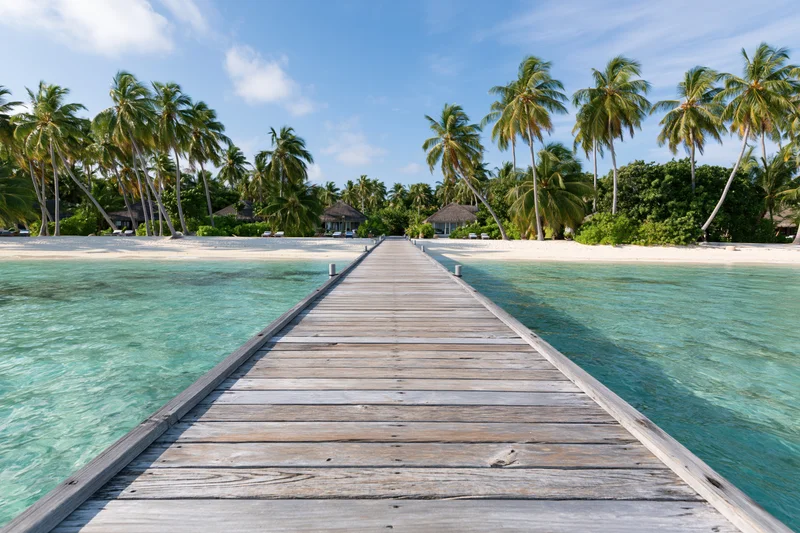 Wooden dock extending over turquoise water toward palm-fringed beach with bungalows