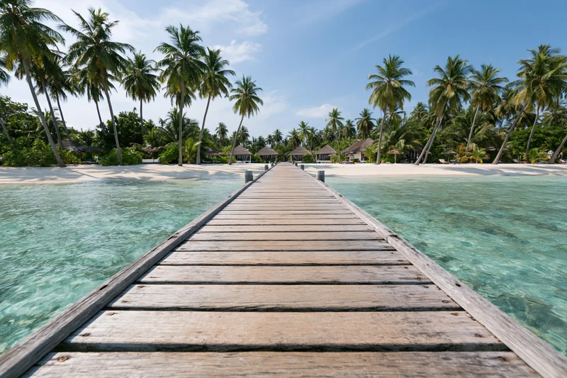 Wooden dock extending over turquoise water toward palm-fringed beach with resort bungalows