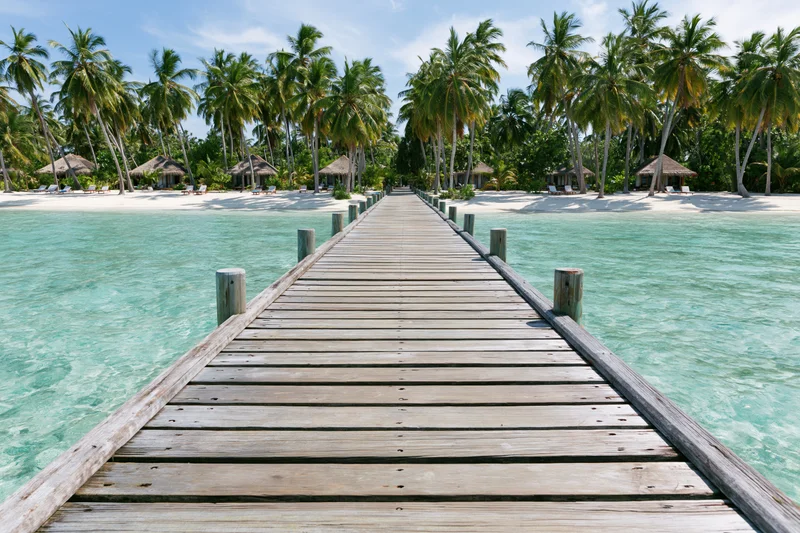 Wooden dock extending over turquoise water toward palm-lined beach with thatched huts