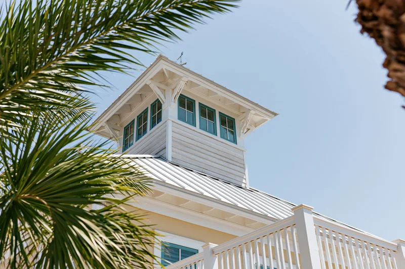 White coastal house with blue windows and palm fronds framing the tower under clear sky