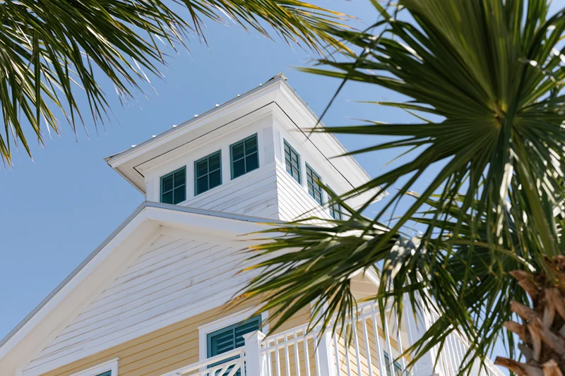 White house with yellow siding and blue shutters framed by palm fronds under a clear blue sky