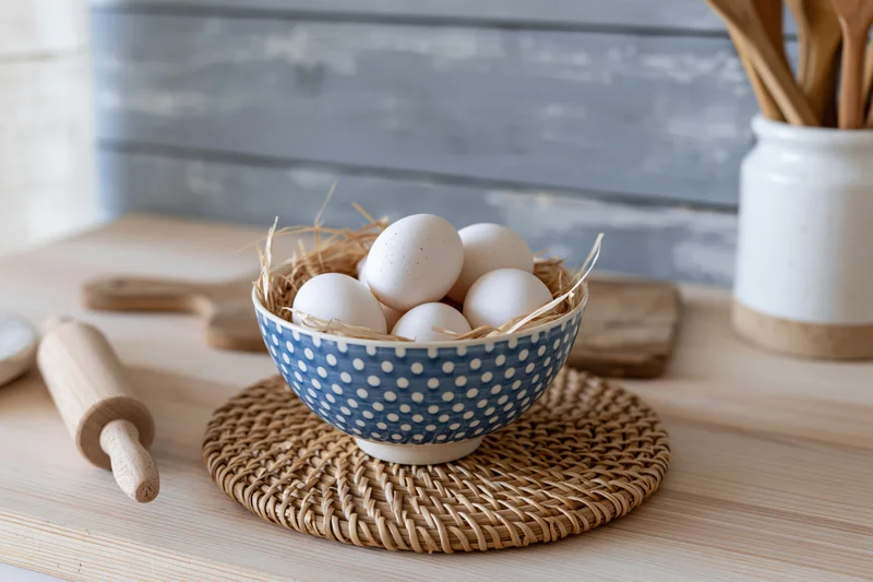 White eggs in a blue polka dot bowl lined with straw on a wooden countertop surrounded by baking tools