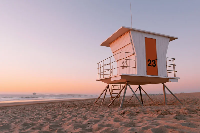 Lifeguard tower on sandy beach at sunset with ocean and pier in distance