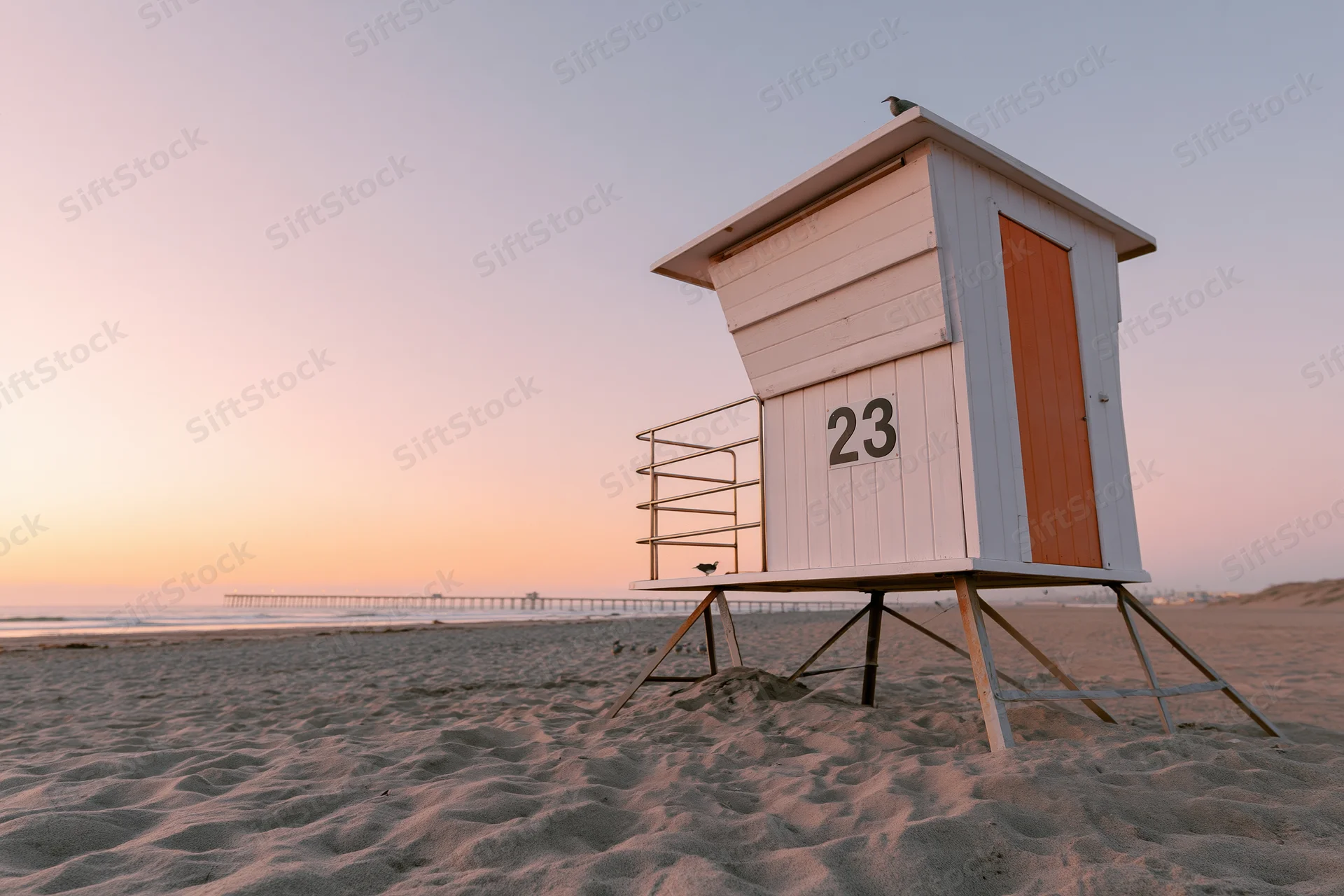 White lifeguard tower with number 23 on sandy beach at sunset near ocean and pier