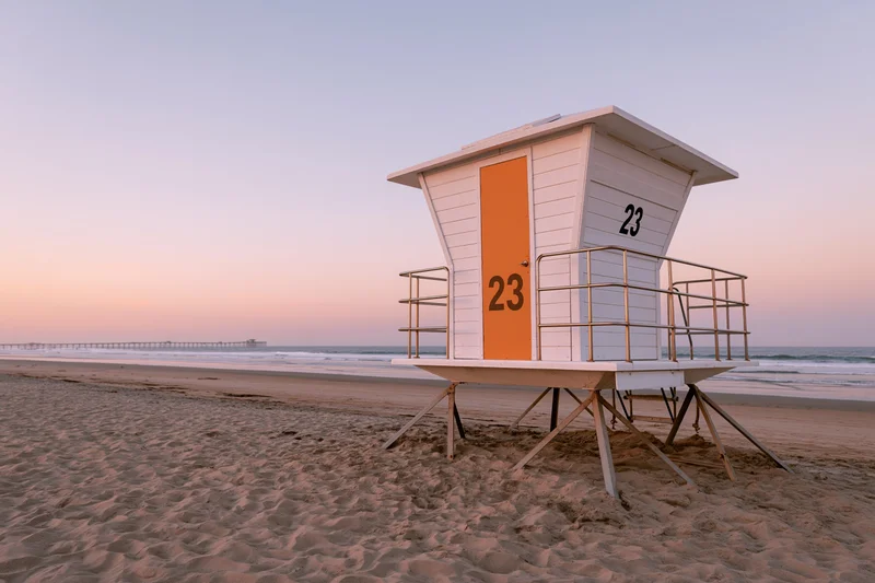 White lifeguard tower with orange door and number 23 on sandy beach at sunset near ocean