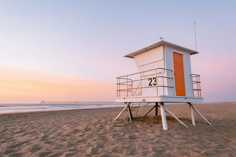 White lifeguard tower with orange door on sandy beach at sunset near ocean pier