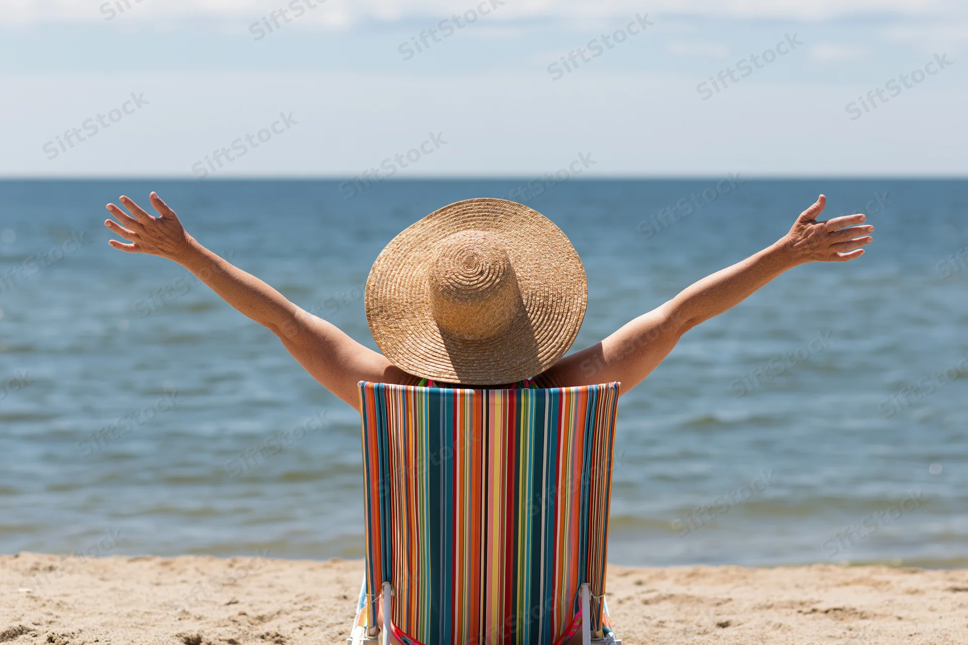 Woman sitting in striped deckchair with arms outstretched facing ocean on sandy beach