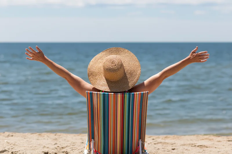 Woman sitting in striped deckchair with arms outstretched facing ocean on sandy beach