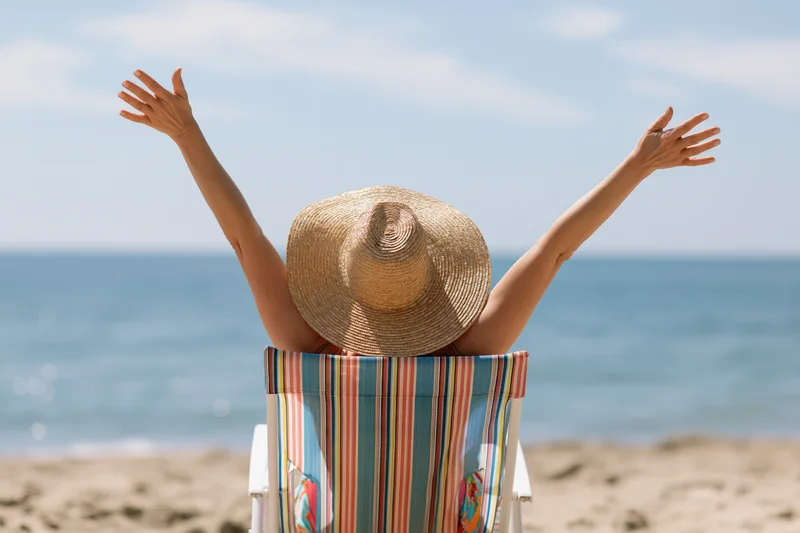 Woman sitting in striped deckchair with arms raised facing ocean on sandy beach
