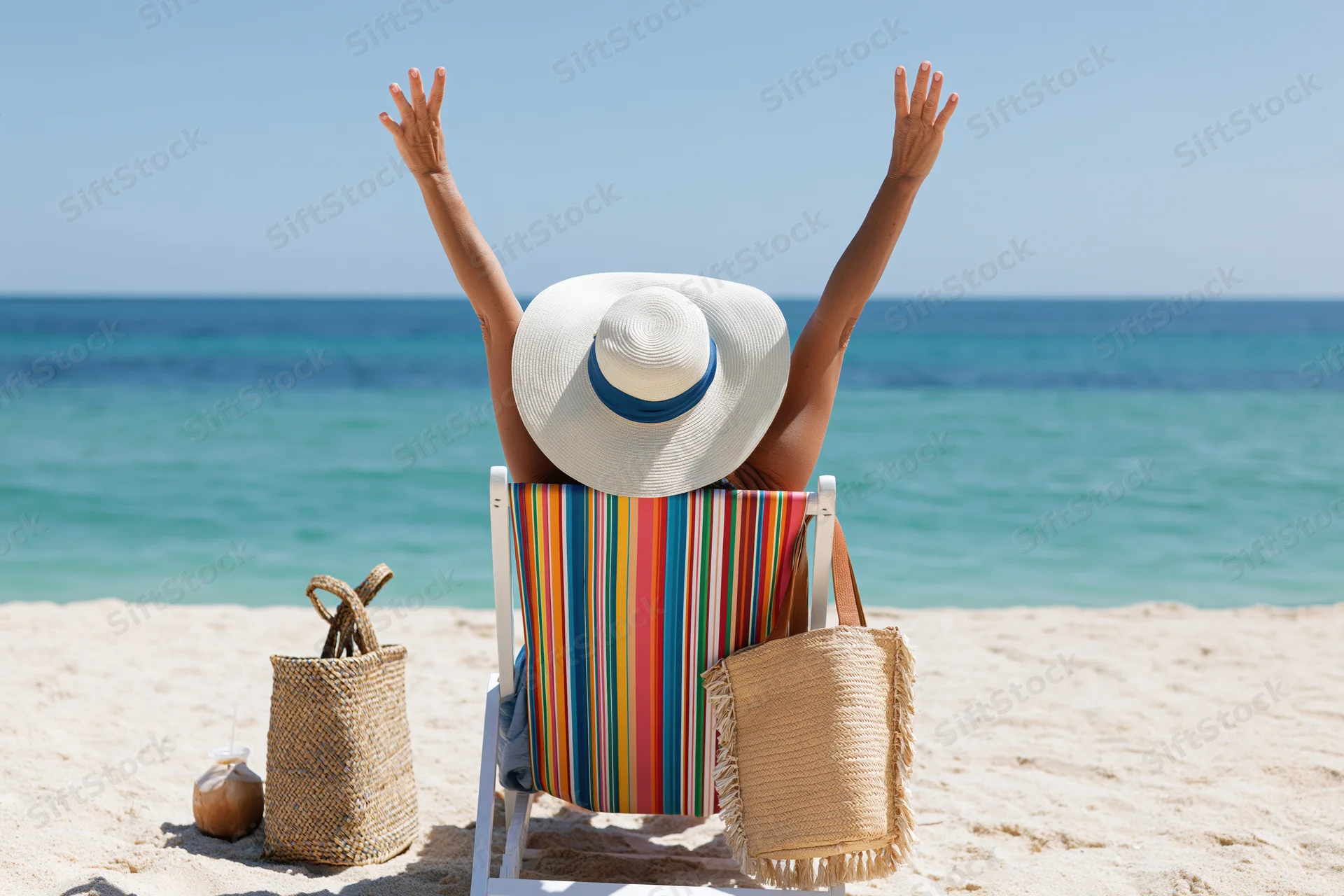 Woman sitting in striped deckchair with arms raised on sandy beach facing ocean
