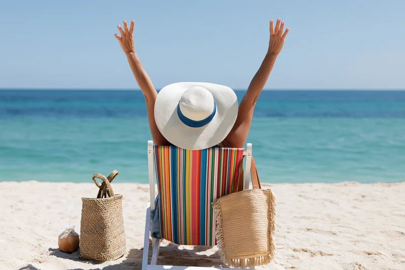 Woman sitting in striped deckchair with arms raised on sandy beach facing ocean
