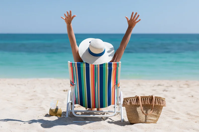 Woman sitting in striped deckchair with arms raised on sandy beach facing ocean