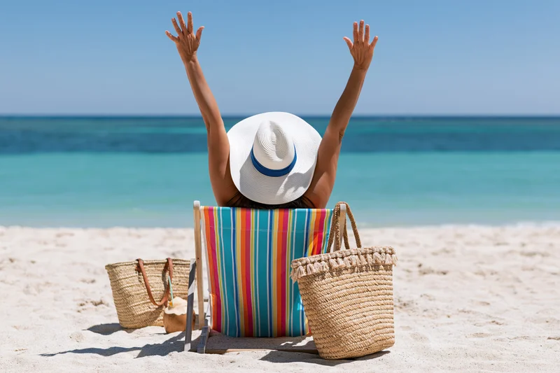 Woman sitting on striped beach chair with arms raised on sandy beach facing ocean