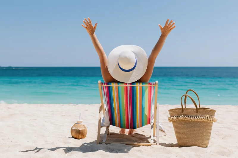 Woman sitting in striped deckchair with arms raised on sandy beach facing ocean