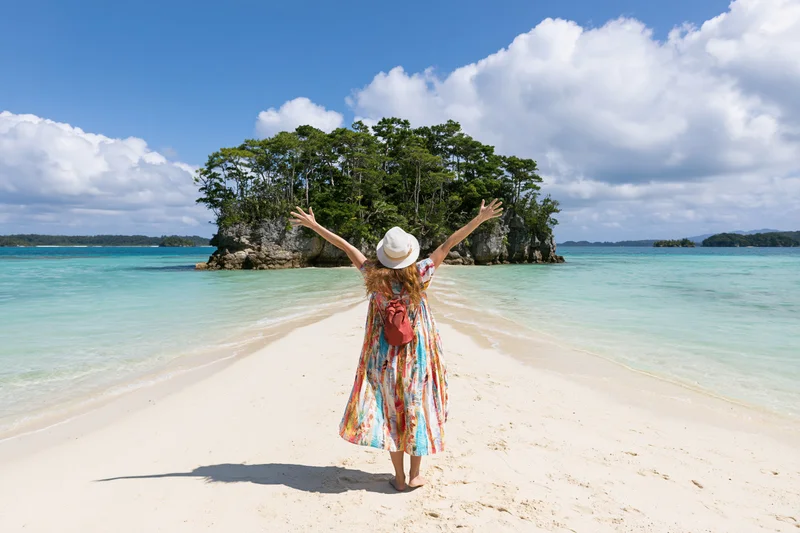 Woman with arms raised standing on sandy beach facing tropical island and turquoise sea