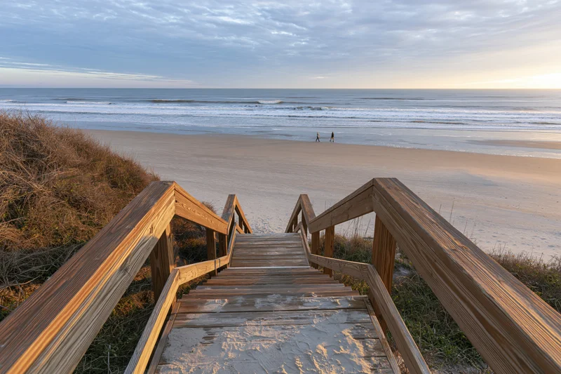 Wooden staircase leading down to sandy beach with ocean waves and distant figures at twilight