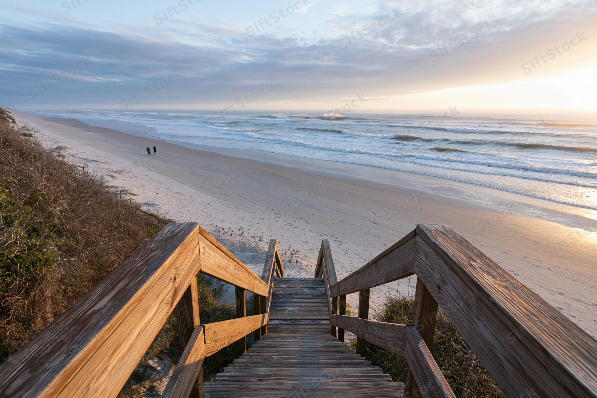 Wooden staircase leading down to sandy beach at sunset with ocean waves and dunes in view