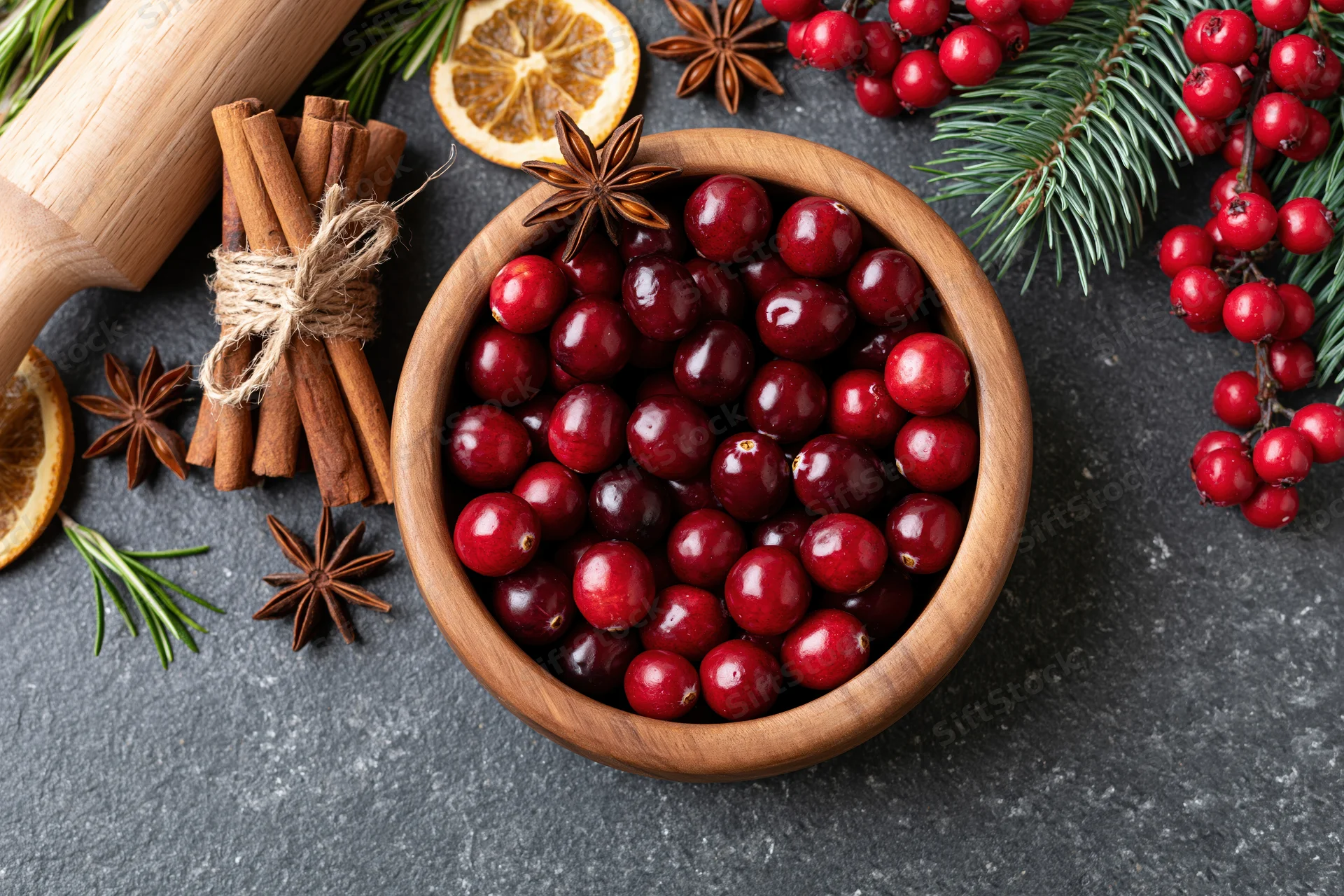 Fresh cranberries in a wooden bowl surrounded by cinnamon sticks, star anise, orange slices, rosemary, and evergreen sprigs on a dark countertop