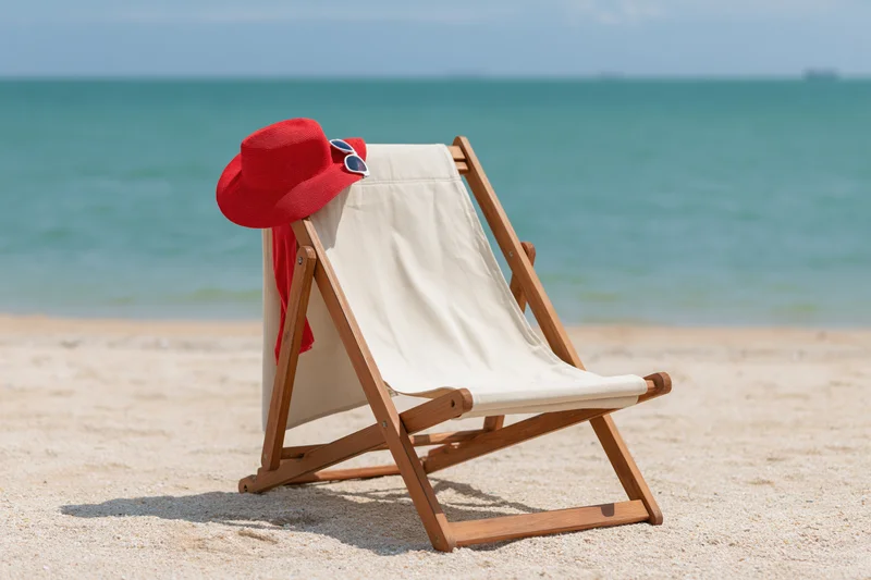 Wooden deckchair with red sunhat and sunglasses on sandy beach by ocean
