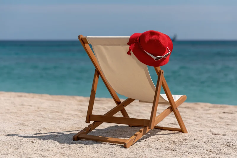 Wooden deckchair with red hat and sunglasses on sandy beach overlooking ocean