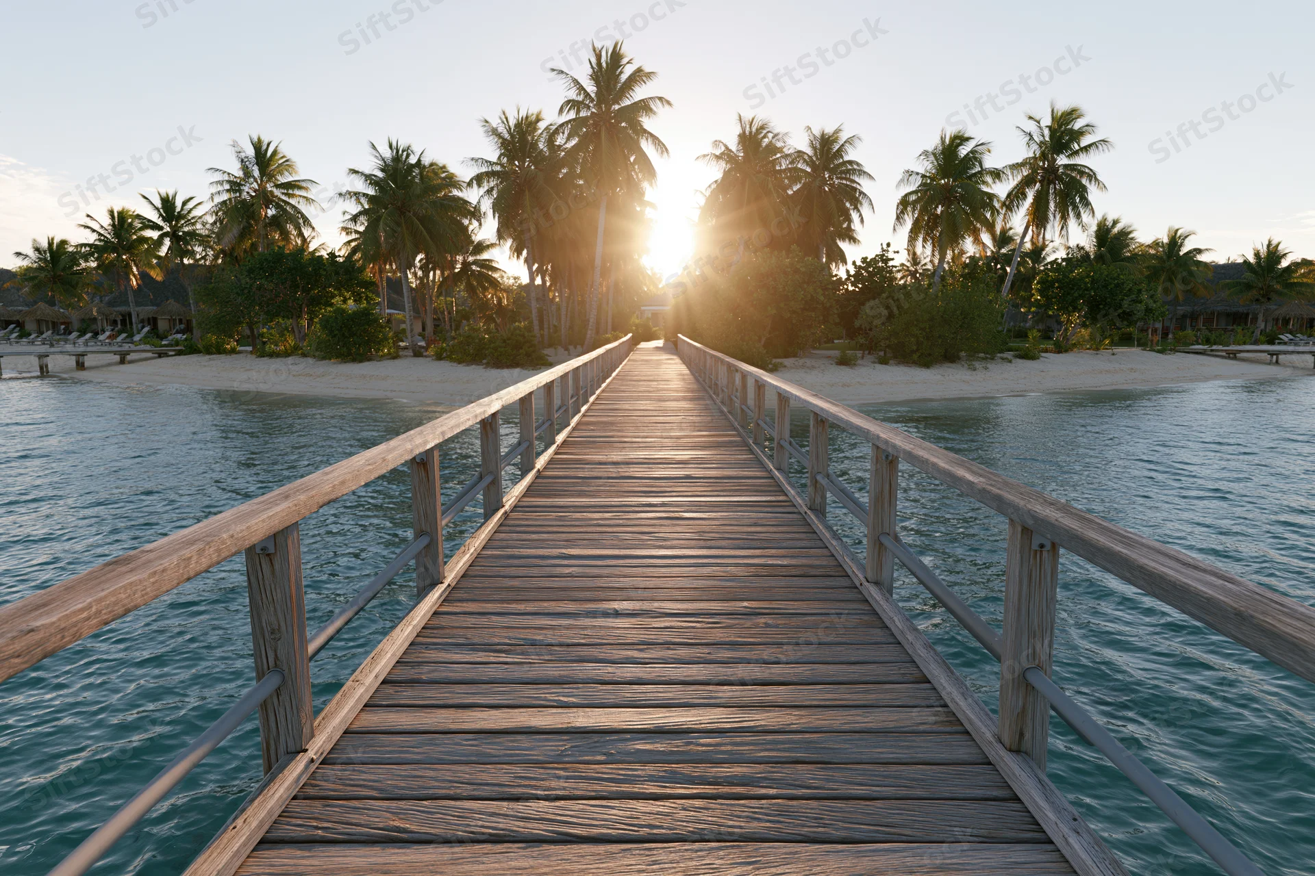 Wooden pier extending over calm ocean toward tropical beach at sunset with palm trees lining the shore