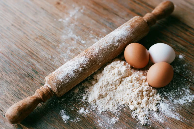 Wooden rolling pin dusted with flour beside eggs and pile of flour on rustic wooden table