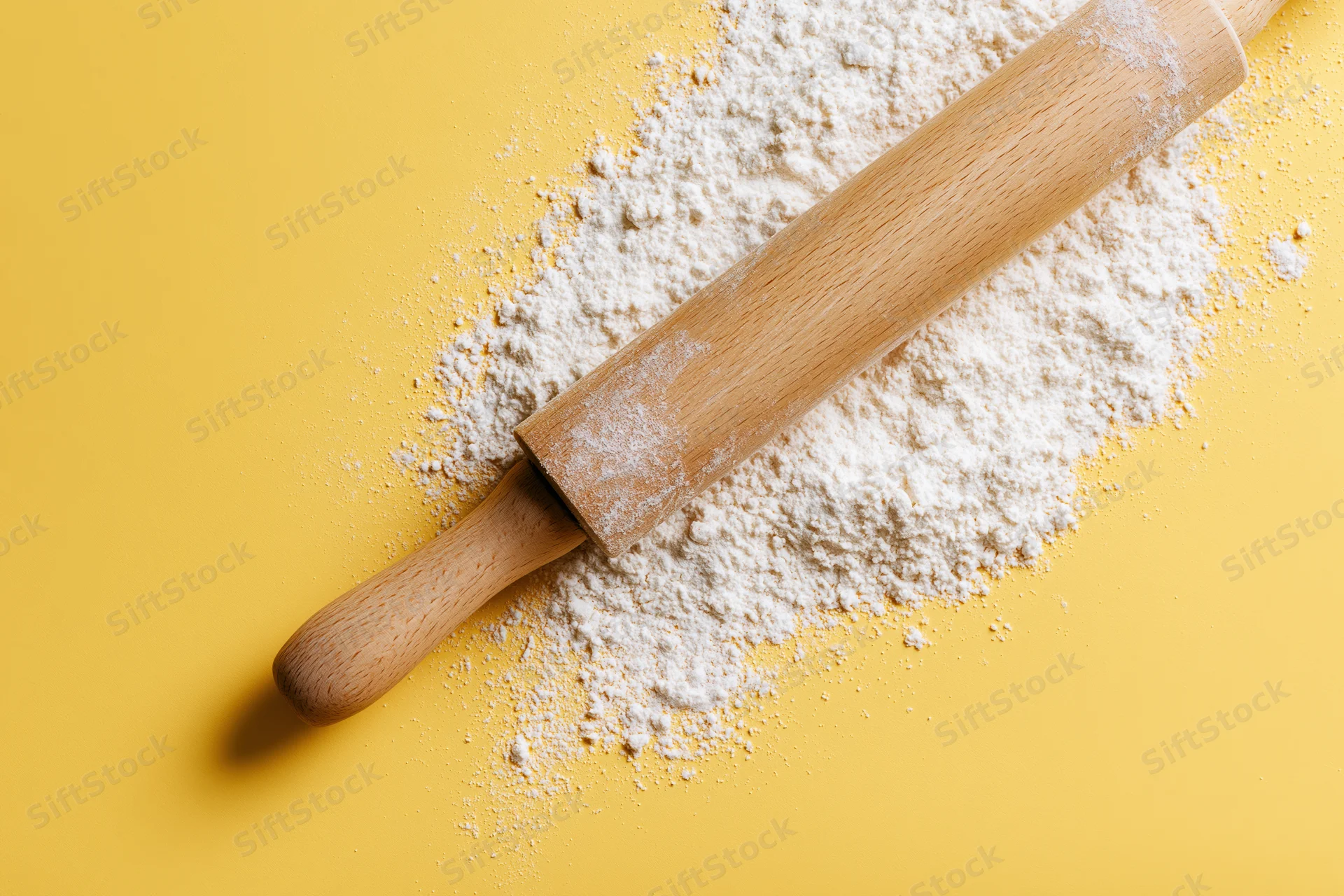 Wooden rolling pin resting on a pile of white flour against a yellow background