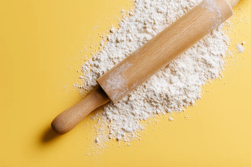 Wooden rolling pin resting on a pile of white flour against a yellow background