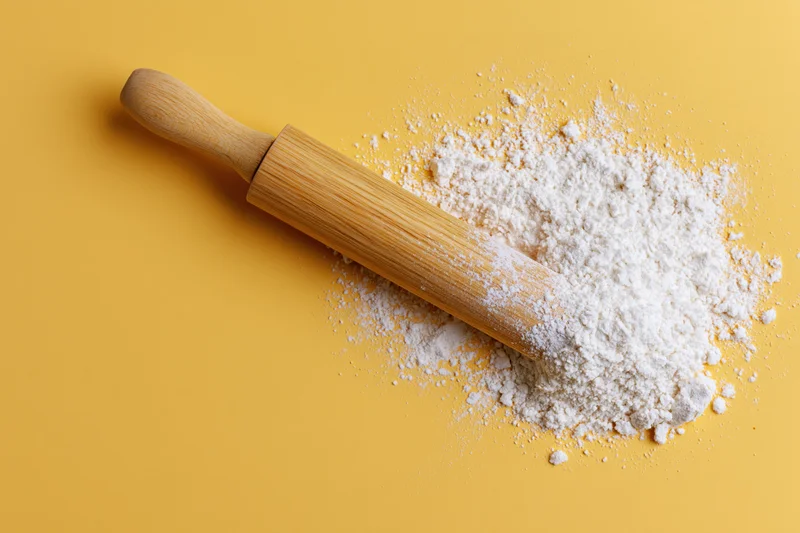 Wooden rolling pin resting on a pile of white flour against a yellow background