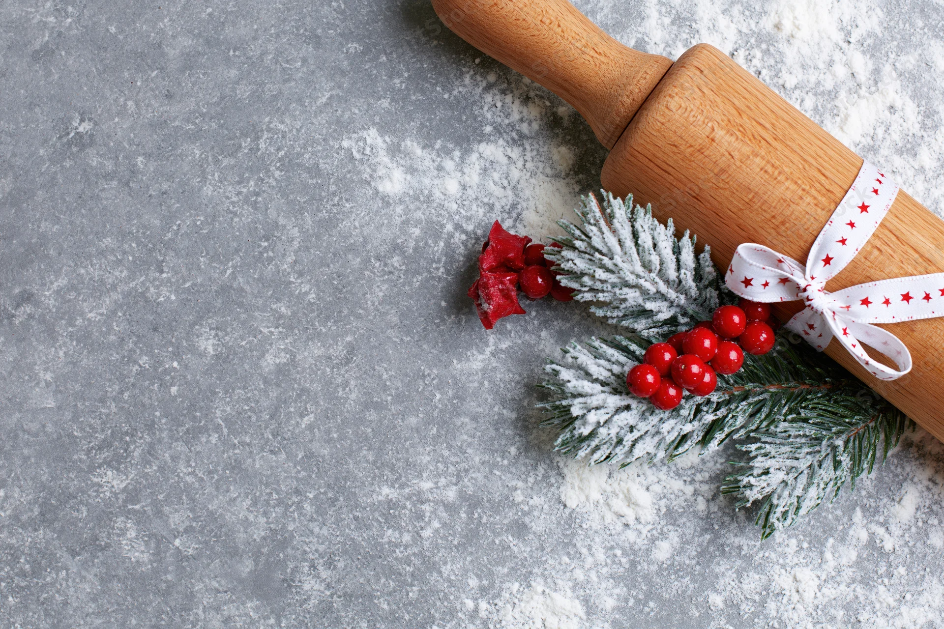 Wooden rolling pin decorated with pine sprig, red berries and ribbon, resting on floured gray countertop