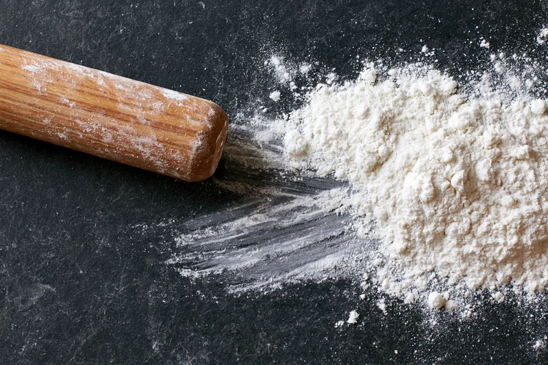 Wooden rolling pin resting beside a pile of flour on a dark countertop