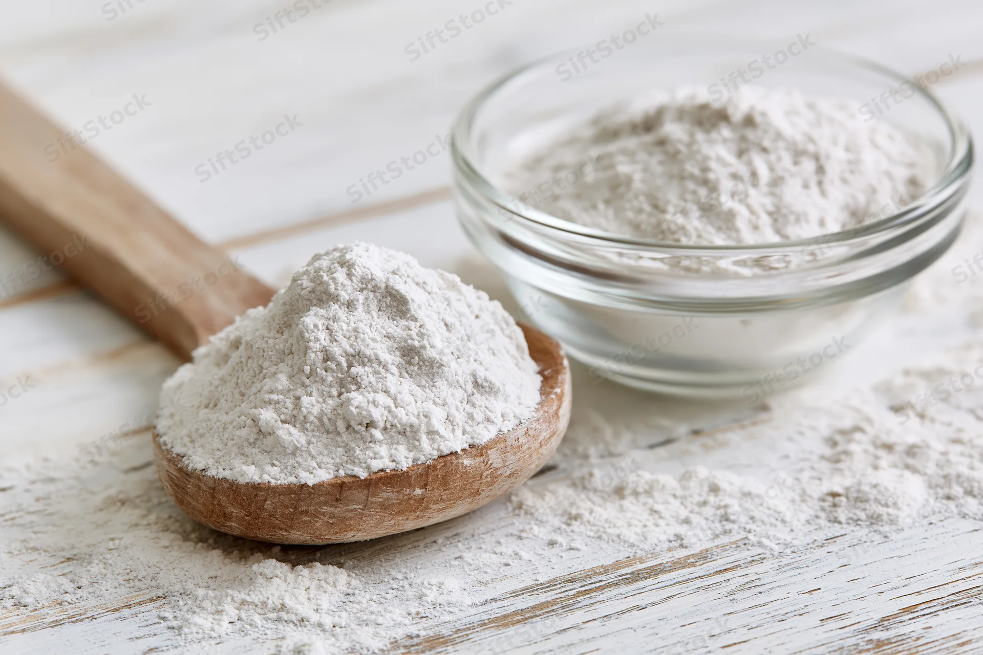 Flour piled on a wooden spoon and in a glass bowl on a white wooden surface
