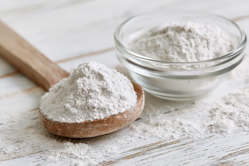 Flour piled on a wooden spoon and in a glass bowl on a white wooden surface