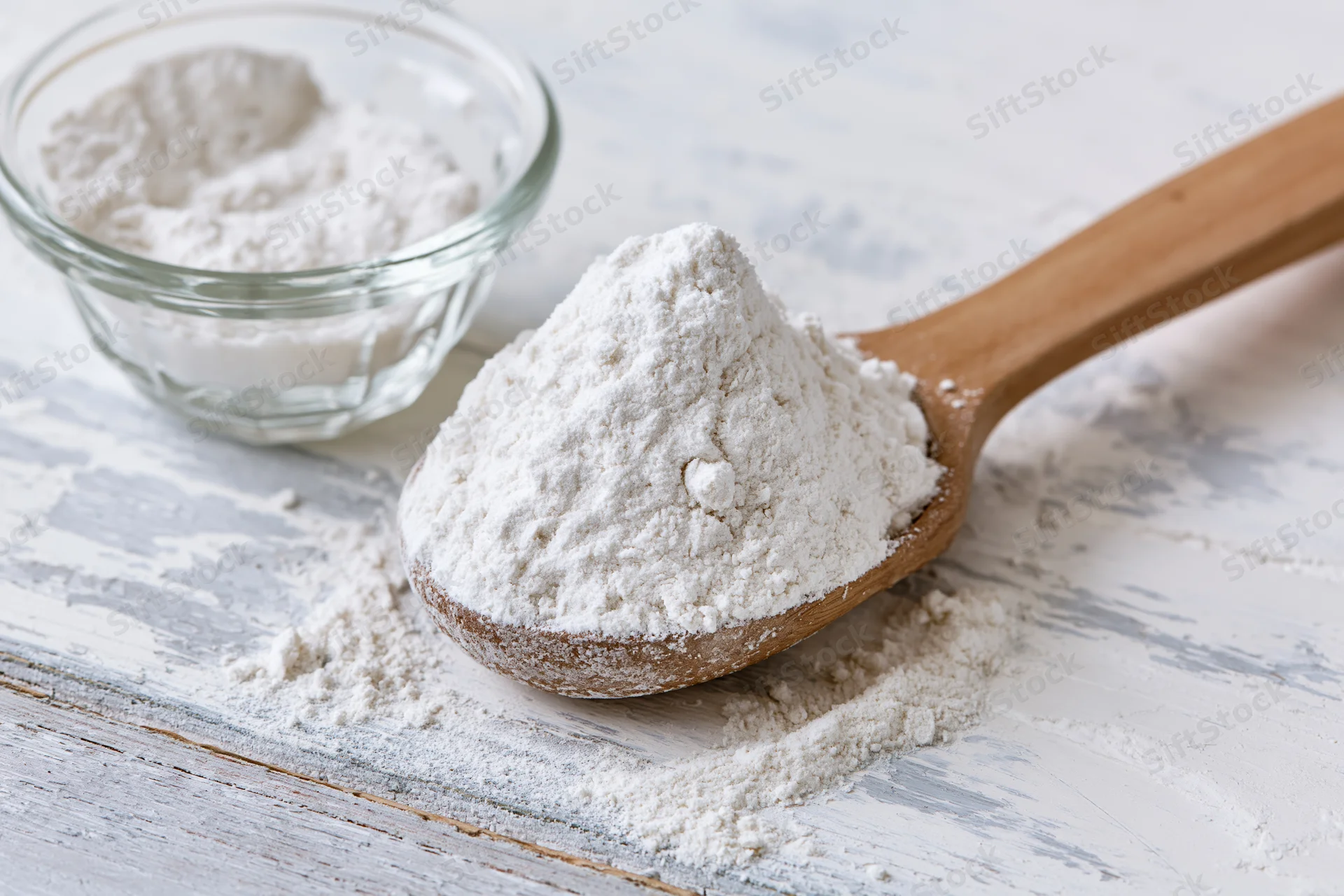 Wooden spoon heaped with flour resting on a distressed white wooden surface beside a glass bowl of flour