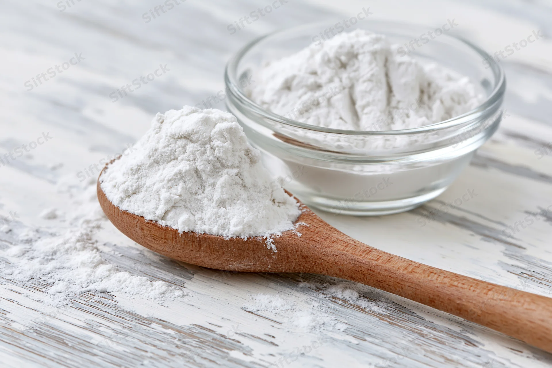 Wooden spoon filled with flour resting beside a glass bowl on a white wooden surface