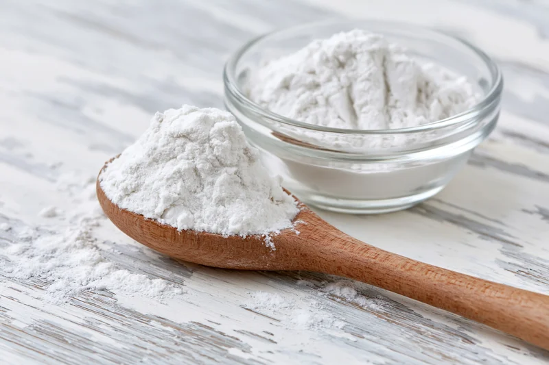 Wooden spoon filled with flour resting beside a glass bowl on a white wooden surface
