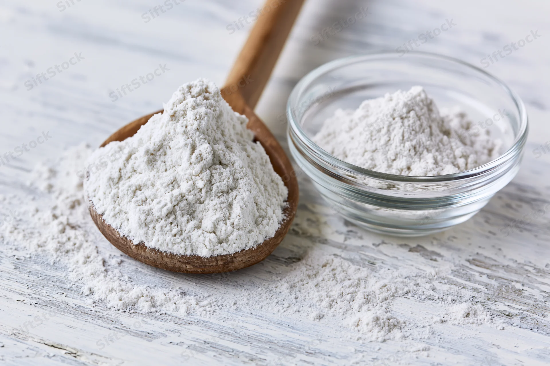 Flour in a wooden spoon and glass bowl on a rustic white table
