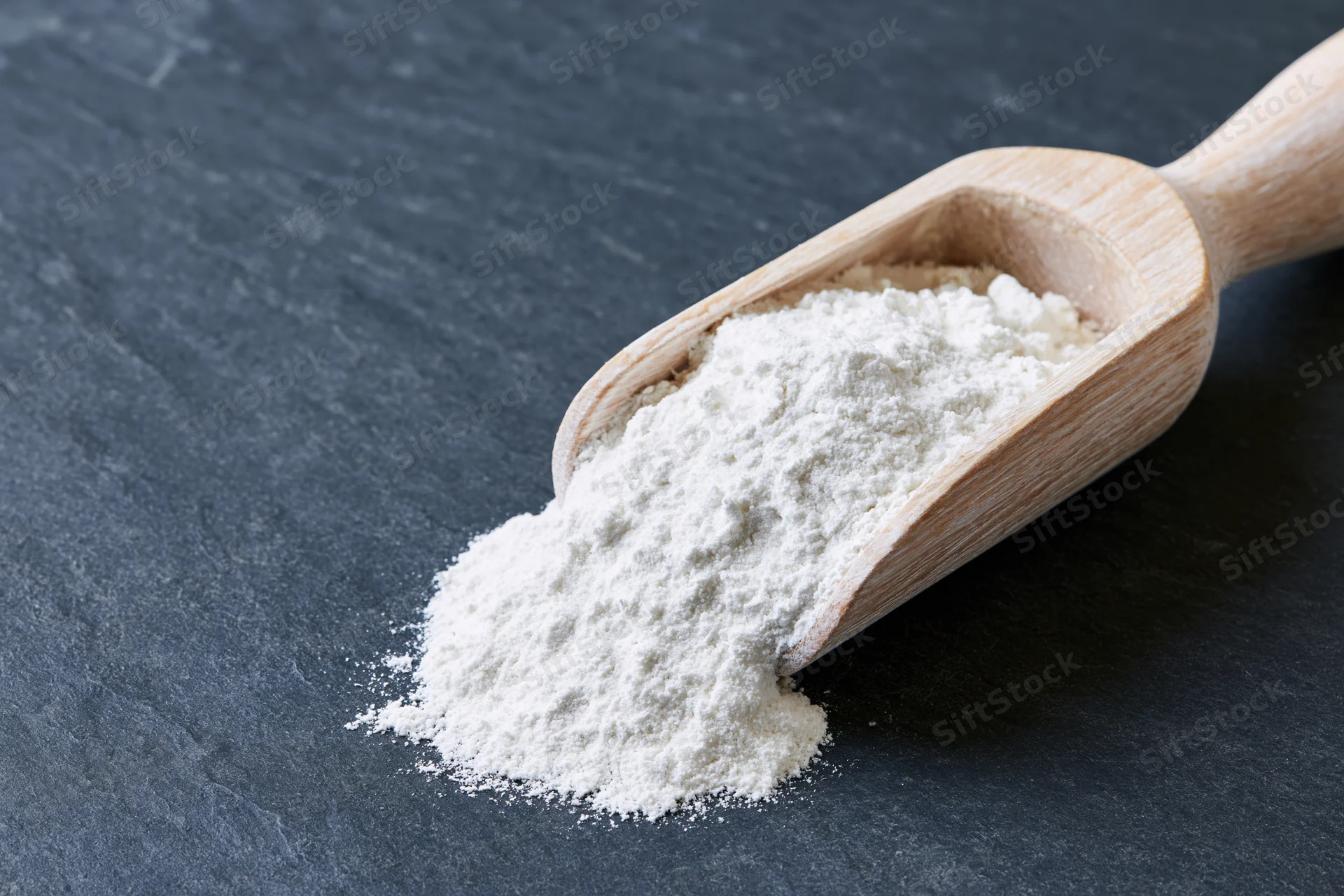 White flour spilling from a wooden scoop on a dark slate surface