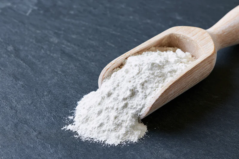 White flour spilling from a wooden scoop on a dark slate surface