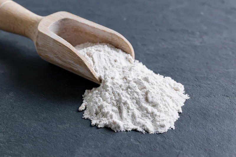 Wooden scoop holding flour spilled onto dark slate surface