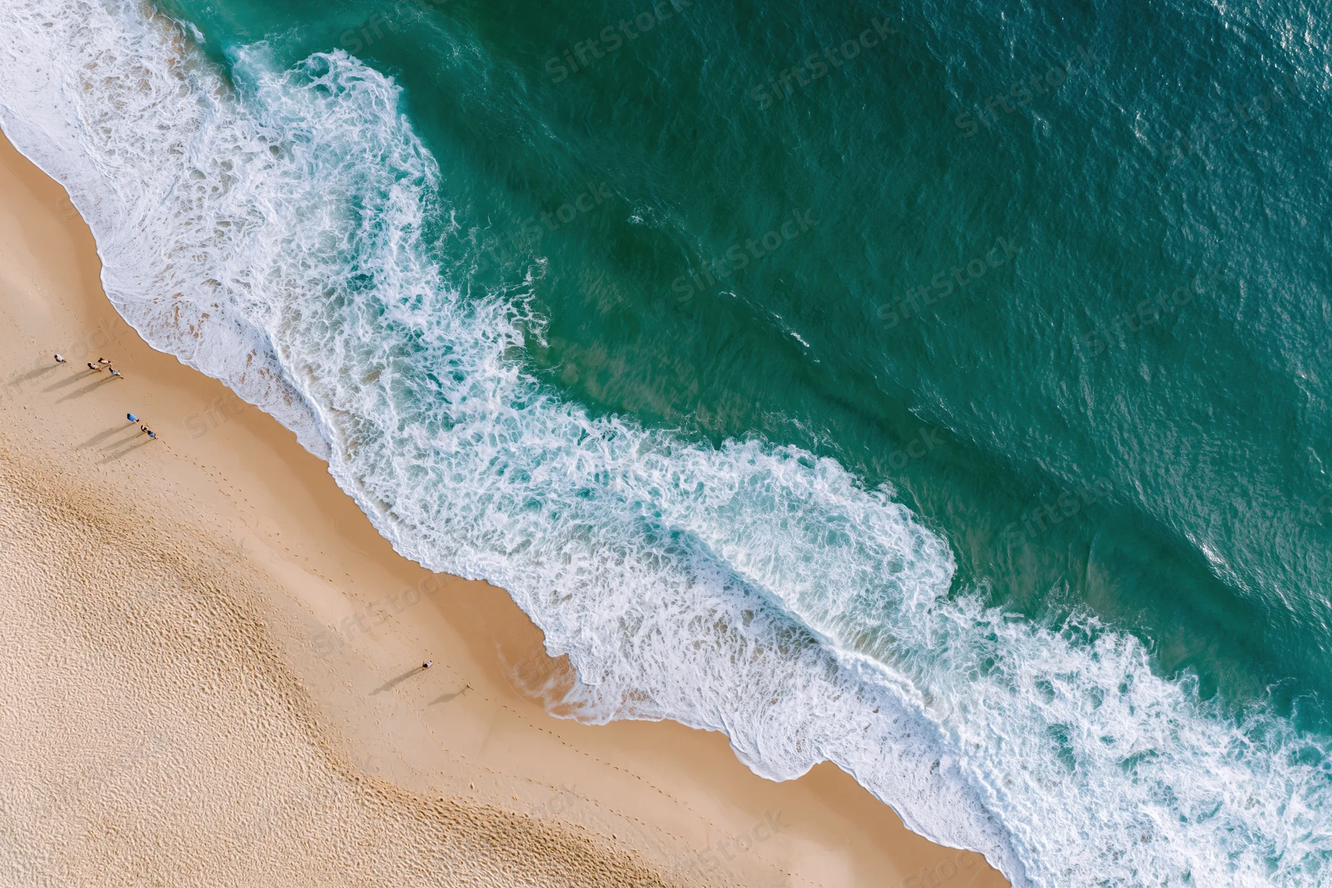 Aerial view of turquoise ocean waves crashing onto sandy beach with small distant people along shoreline
