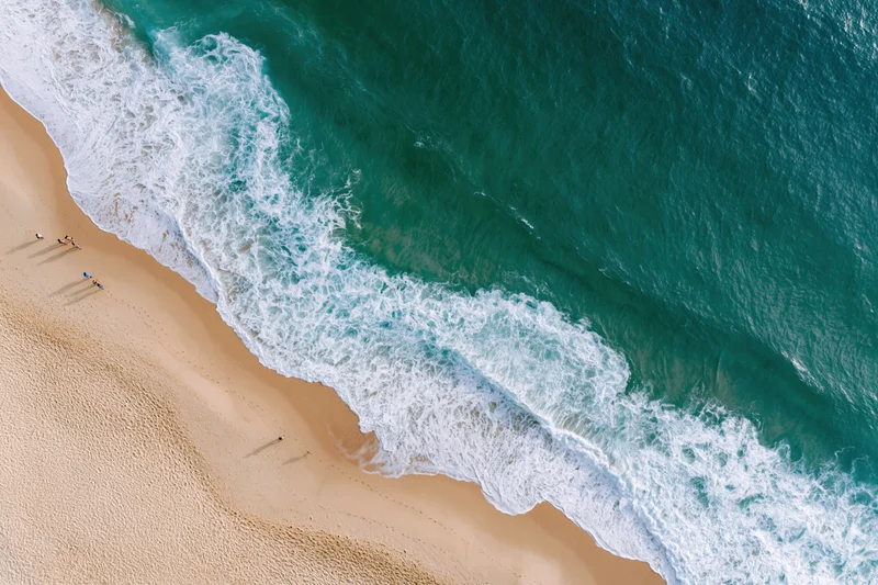 Aerial view of turquoise ocean waves crashing onto sandy beach with small distant people along shoreline