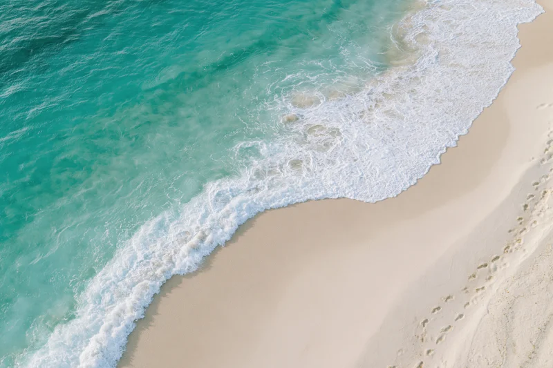 Turquoise waves breaking onto a sandy beach from above, leaving footprints in the sand