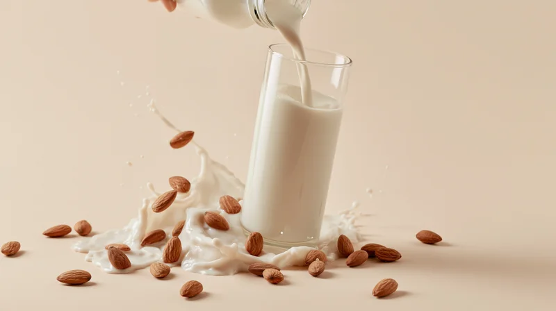 Almond milk being poured into a glass with scattered almonds and splashes on a beige surface