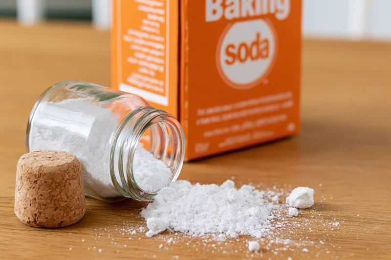 Spilled baking soda powder from a glass jar on a wooden kitchen table with an orange box in the background