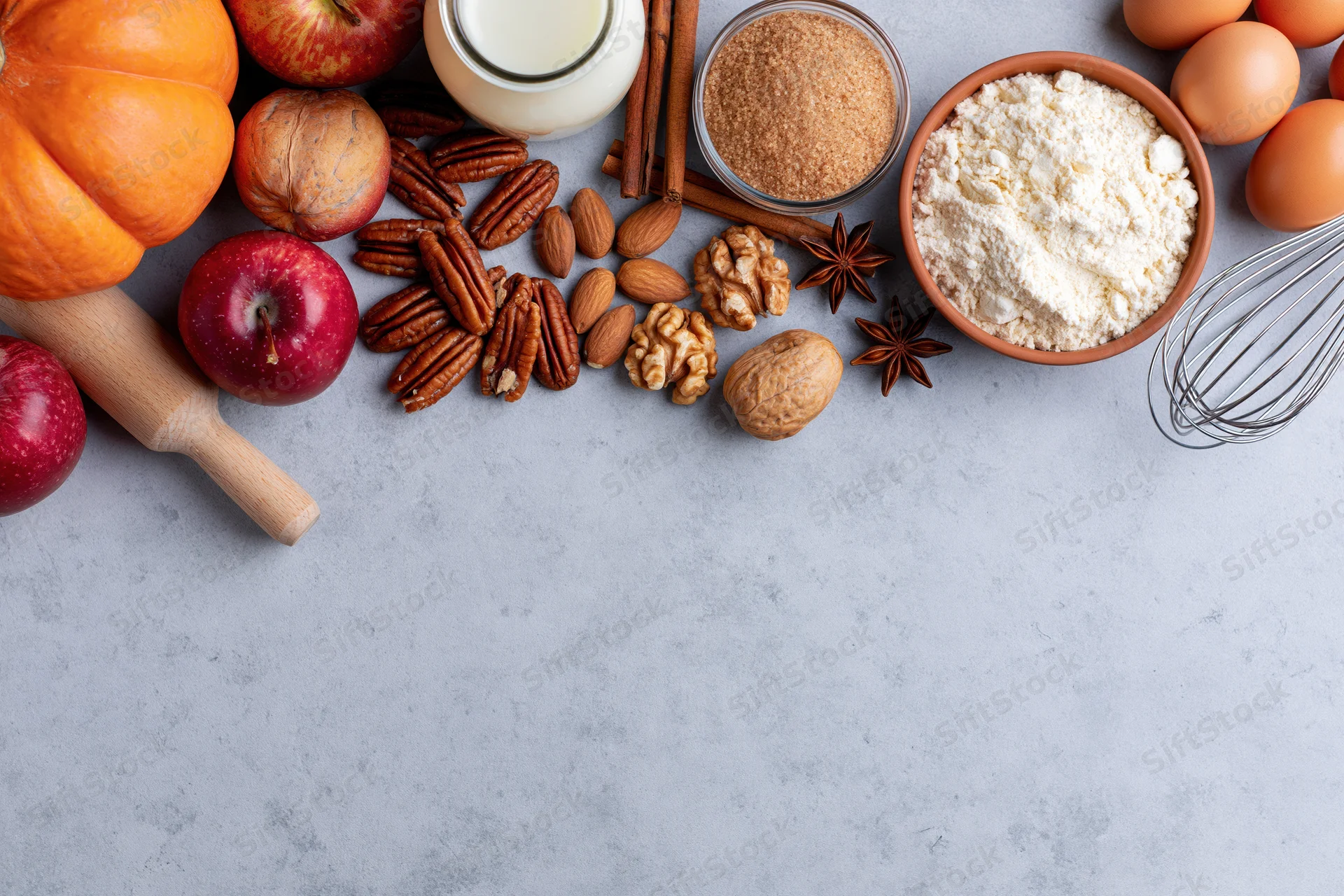 Pumpkin, apples, flour, eggs, milk, nuts, spices, rolling pin and whisk arranged on gray countertop for baking preparation