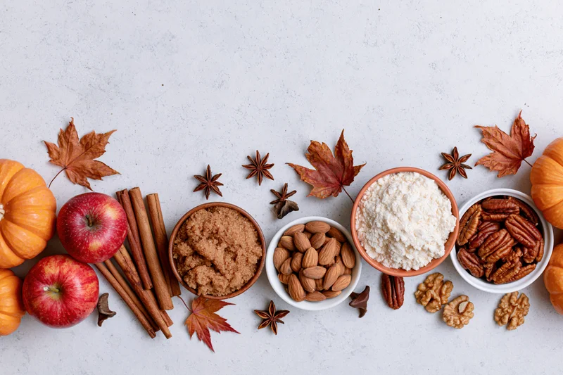 Autumn ingredients arranged in a flat lay on a white surface with pumpkins, apples, cinnamon, nuts, and spices