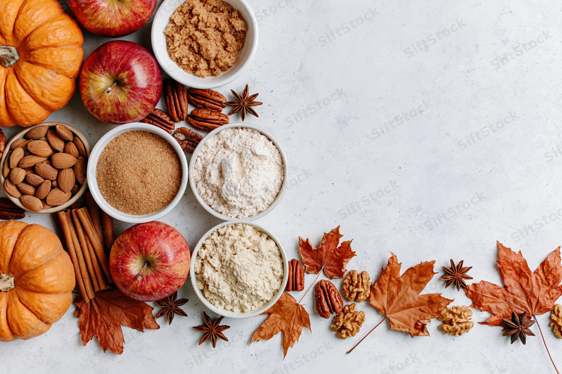 Autumn ingredients including pumpkins, apples, nuts, spices, and sugars arranged on a white countertop with fall leaves and copyspace