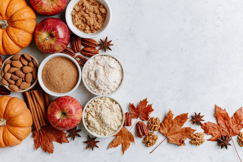 Autumn ingredients including pumpkins, apples, nuts, spices, and sugars arranged on a white countertop with fall leaves and copyspace